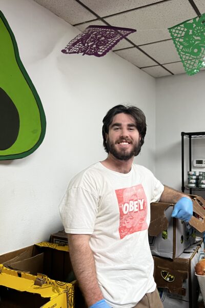A smiling male volunteer wearing gloves standing next to food donation boxes in a decorated room.