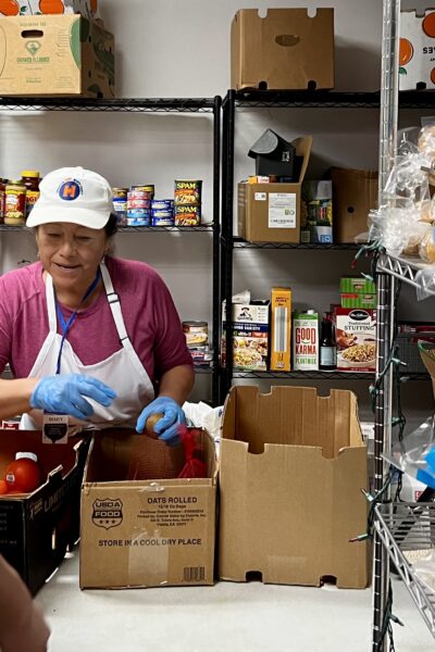 A volunteer with gloves and a cap sorting food donations into cardboard boxes in the pantry.