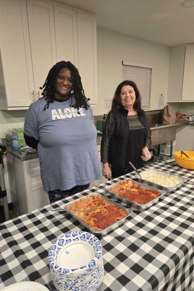 Two smiling volunteers standing behind a table with hot meals and pasta prepared for shelter residents.