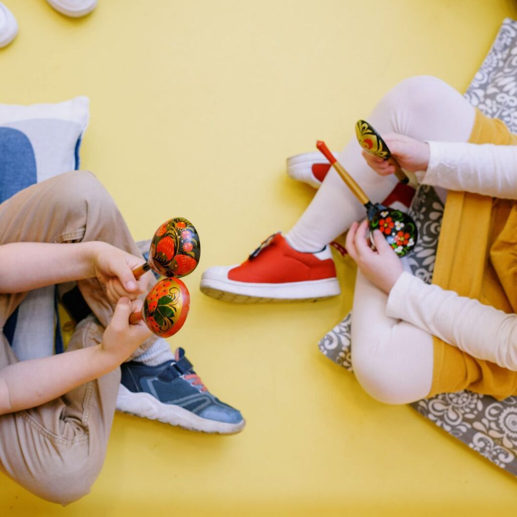 Top-down view of two young children sitting on a bright yellow floor and playing with colorful wooden spoons.
