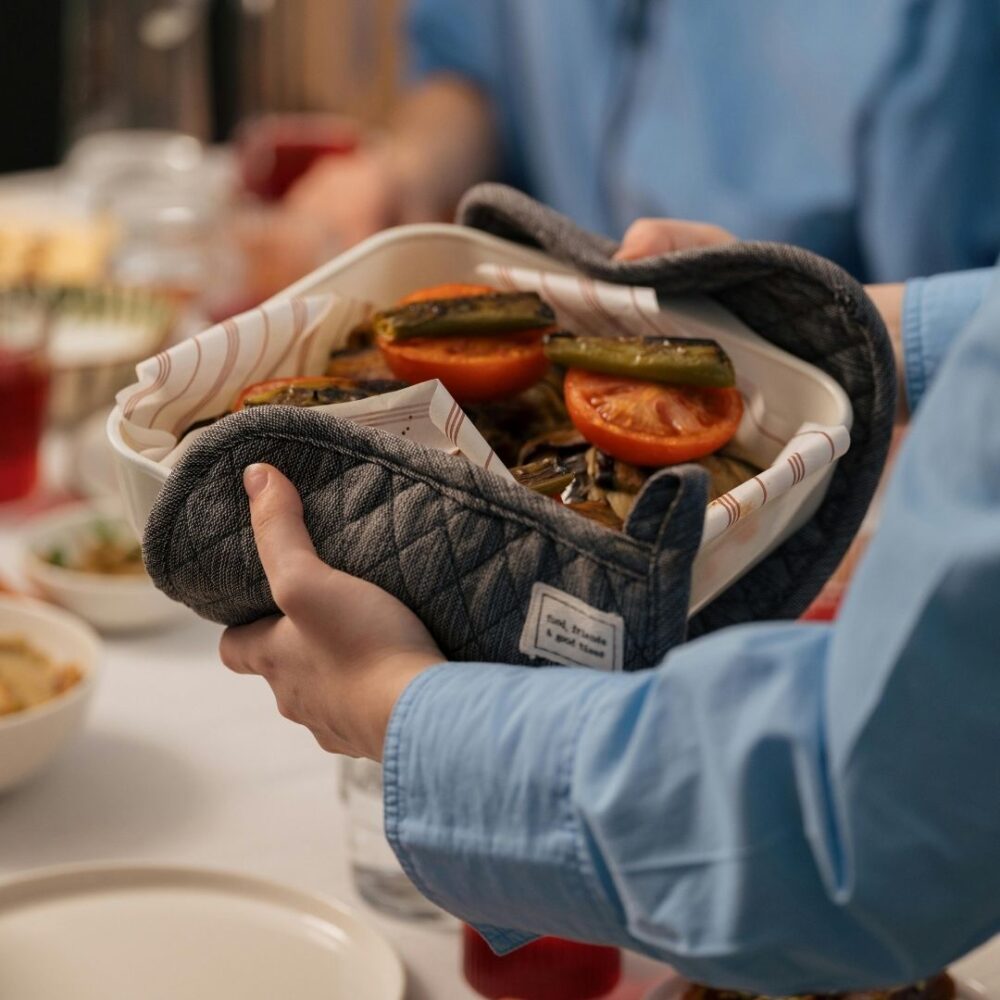 Close-up of a person using oven mitts to serve a hot dish of roasted vegetables at a dining table.