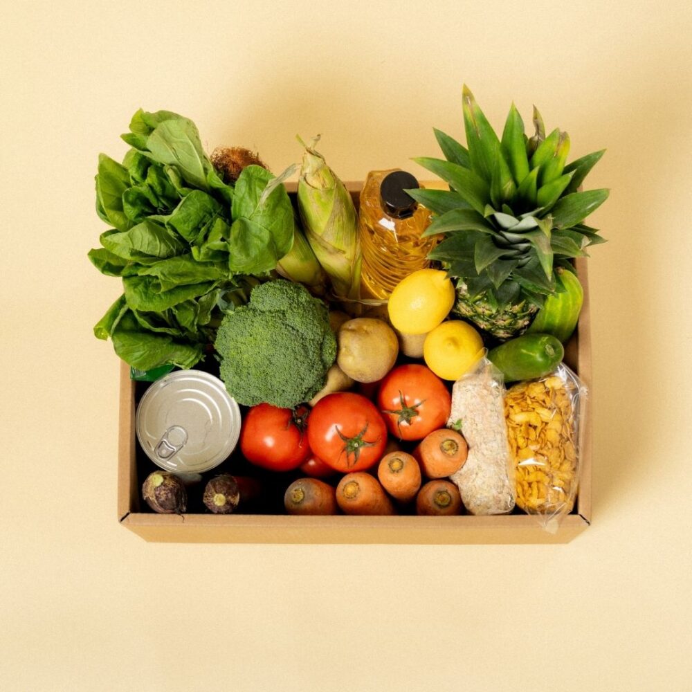 Top-down view of a cardboard donation box packed with fresh produce, cooking oil, canned goods, and pantry staples.