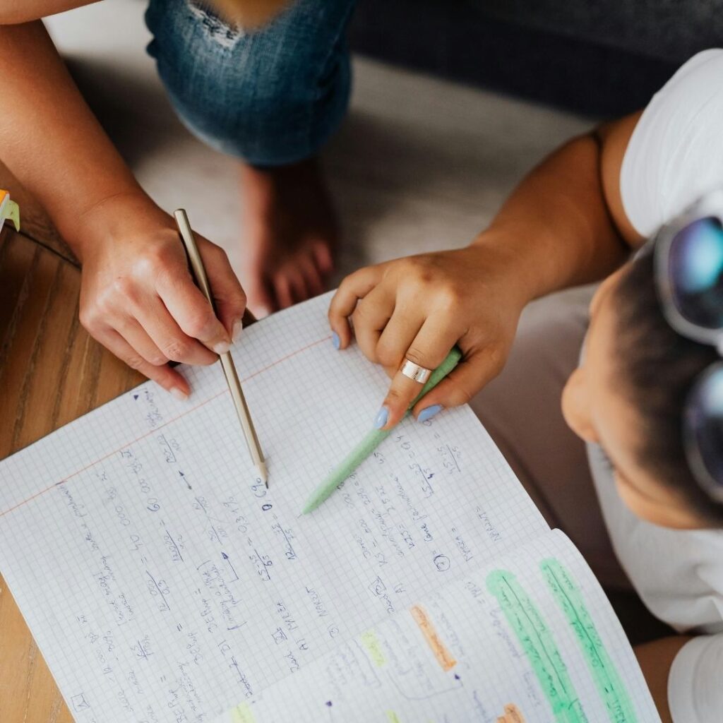 Top-down view of two people studying together, pointing at notes and math equations in a grid notebook with pens.