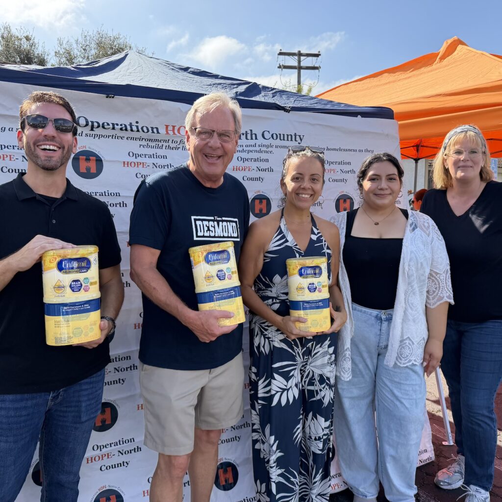 Group of smiling volunteers in front of an event tent, holding cans of donated baby formula.
